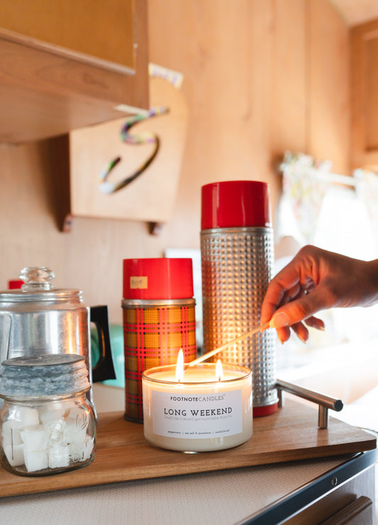 Long Weekend Candle from Footnote Candles. Candle is sitting with vintage camping items, being lit with long stick match.