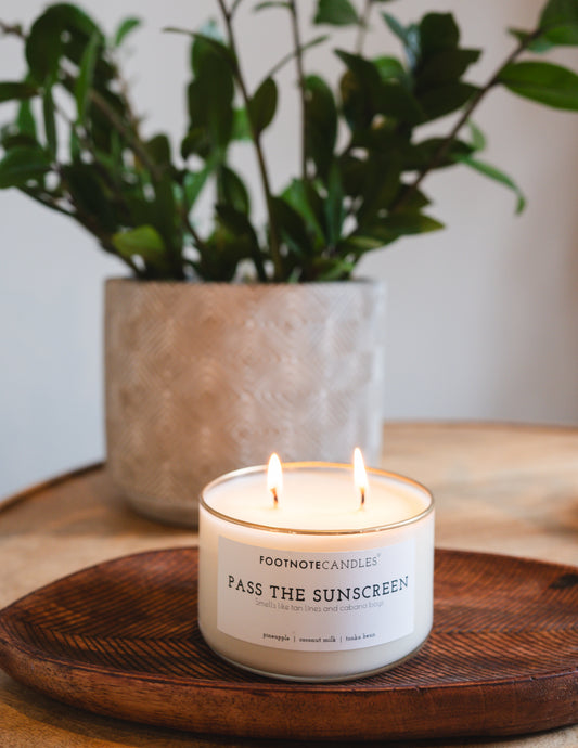 Pass the Sunscreen Candle from Footnote Candles. Candle is sitting on textures wood tray with houseplant in background.